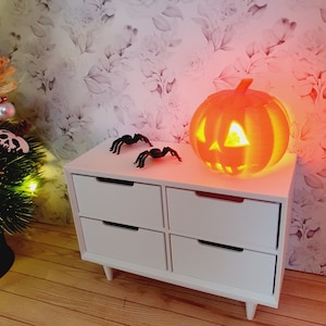 Può includere: Una cassettiera bianca con quattro cassetti e figurine di ragni neri in cima. Una zucca arancione incandescente si trova sulla cassettiera.