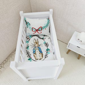 Puede incluir: Cuna de madera blanca con un juego de ropa de cama blanco y verde con un diseño de Peter Rabbit. La ropa de cama incluye una almohada con un lazo rojo y una corona verde.