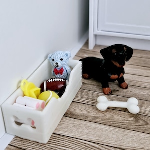 May include: A white wooden toy bin with a bone-shaped cutout on the front. The bin is filled with toys, including a blue teddy bear with a red bow tie, a yellow squeaky toy, a tennis ball, a football, and a white bone. A black dachshund dog toy is sitting on the floor next to the bin.