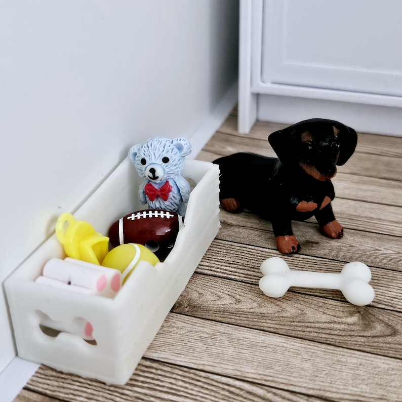 May include: A white wooden toy bin with a bone-shaped cutout on the front. The bin is filled with toys, including a blue teddy bear with a red bow tie, a yellow squeaky toy, a tennis ball, a football, and a white bone. A black dachshund dog toy is sitting on the floor next to the bin.