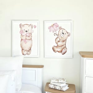 May include: Two rectangular wall art prints featuring watercolour illustrations of light brown teddy bears. The left print shows a bear holding pink hearts, while the right print shows a bear holding a pink flower. Both are in white frames.
