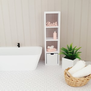 May include: White bathroom shelf unit with three shelves and a drawer. The top shelf has the word "Bath" in pink lettering. The middle shelf has a pink bottle and the bottom shelf has a pink box. A green plant is in a white pot on the right side of the shelf unit.