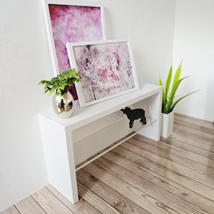 Puede incluir: Una consola blanca con dos impresiones artísticas abstractas enmarcadas. Un jarrón plateado con vegetación y una silueta negra de animal están sobre la mesa. Un jarrón blanco alto con plantas verdes está a la derecha.