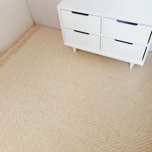 May include: A light beige woven rug with a herringbone pattern. The rug has a fringed edge and is next to a white four-drawer chest of drawers with black drawer pulls. The rug is a neutral colour and would complement many decor styles.