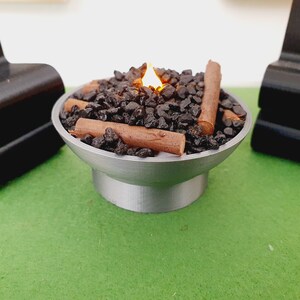K&ouml;nnte beinhalten: Ein Miniatur-Tischfeuer mit einer Flamme, schwarzen Steinen und braunen Holzscheiten. Der Feuerplatz ist aus Metall und hat eine runde Basis.