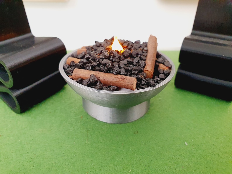 K&ouml;nnte beinhalten: Ein Miniatur-Tischfeuer mit einer Flamme, schwarzen Steinen und braunen Holzscheiten. Der Feuerplatz ist aus Metall und hat eine runde Basis.