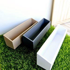 Puede incluir: Tres jardineras rectangulares hechas de diferentes materiales. Las jardineras son beige, gris y blanca. Las cajas están sentadas en un lecho de hierba verde.