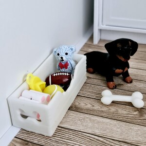 May include: A white wooden toy box with a cutout of a bone on the front. The box is filled with toys, including a teddy bear, a football, a yellow ball, and a whistle. A black and brown dachshund dog toy is sitting next to the box, and a white bone toy is on the floor.