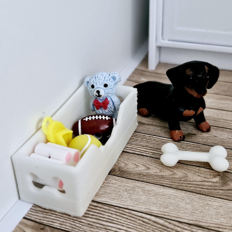 May include: A white wooden toy box with a cutout of a bone on the front. The box is filled with toys, including a teddy bear, a football, a yellow ball, and a whistle. A black and brown dachshund dog toy is sitting next to the box, and a white bone toy is on the floor.