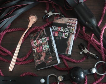 Può includere: Due libri in bianco e nero con il titolo "Bedside Table" sulla copertina. I libri sono su una corda rossa e una frusta di cuoio nero. I libri fanno parte di una piccola zine di galleria.