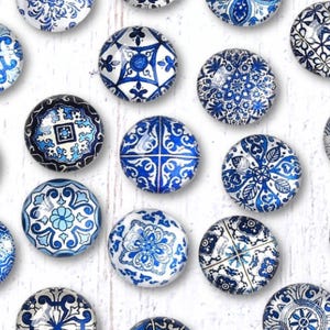 May include: A collection of twenty-one round glass magnets with intricate blue and white patterns. The designs vary, featuring floral, geometric, and abstract motifs. The magnets are arranged on a white, textured surface.