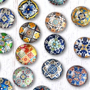 May include: A collection of twenty-one round glass cabochons with colorful, intricate designs. The cabochons feature floral, geometric, and mosaic patterns in shades of blue, yellow, orange, and green. They are arranged on a white wooden surface.