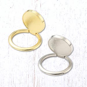 May include: Two metal ring holders with hinged, round tops. One is gold-toned, the other silver-toned. The rings are open, and the tops are slightly ajar, revealing the interior. The background is a white, textured surface.