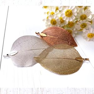 May include: Three decorative leaf-shaped hair clips in silver, gold, and copper tones. The clips are arranged on a white surface with a bouquet of white and yellow daisies in the background. The leaves have detailed vein patterns.