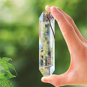 May include: A clear, faceted crystal wand held against a blurred green background. The crystal is rectangular with pointed ends and reflects multiple colors. A thin, dark cord is attached to the top of the crystal.