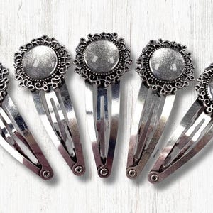 May include: Five silver-toned hair barrettes with ornate, filigree-style frames surrounding a circular, blank center. The barrettes are arranged in a row, showcasing their decorative design and potential for customization.