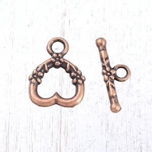 May include: A copper-colored heart-shaped toggle clasp with floral detailing and a matching bar. The clasp features a loop at the top and a decorative floral design around the heart shape. The bar has a loop and floral accents.