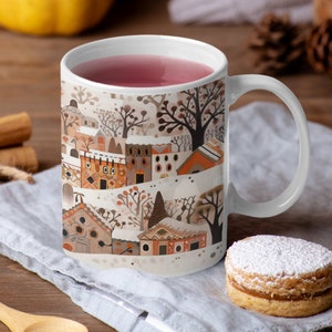 May include: White ceramic mug with a colorful illustration of a winter village scene. The mug is filled with pink liquid and has a white handle.