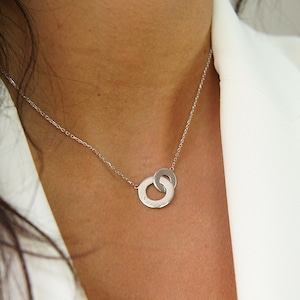 Puede incluir: Un collar de plata con una cadena delicada y un colgante con dos círculos entrelazados.