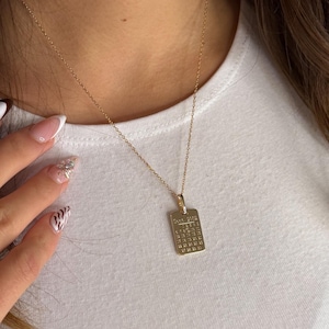 Könnte beinhalten: Eine goldfarbene Halskette mit einem rechteckigen Anhänger. Der Anhänger zeigt ein Kalenderdesign mit dem Datum September 2003. Die Halskette wird auf einem weißen T-Shirt getragen.