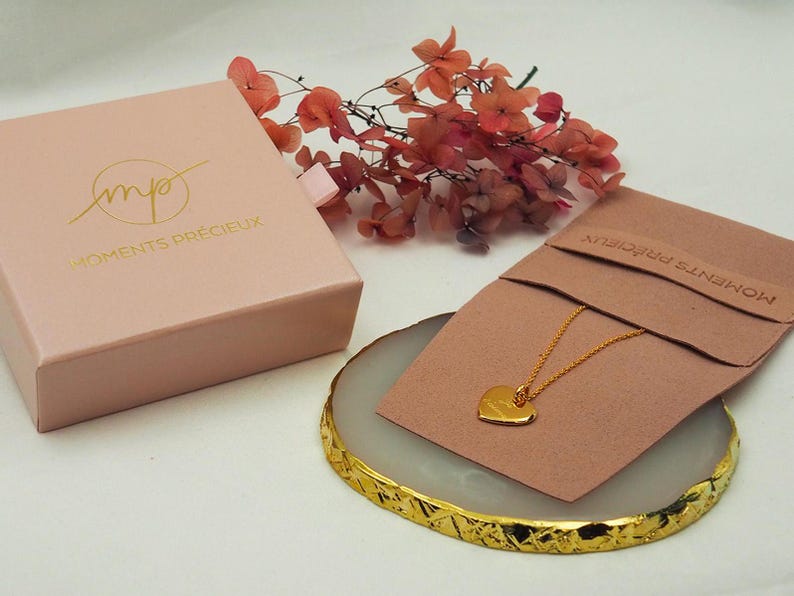 Puede incluir: Un collar de coraz&oacute;n de oro en una pantalla rosa y dorada con el texto "Moments Pr&eacute;cieux" en la caja y la bolsa.