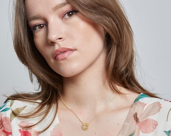 Può includere: Una donna indossa una blusa con stampa floreale e una collana d'oro con un piccolo ciondolo rotondo.