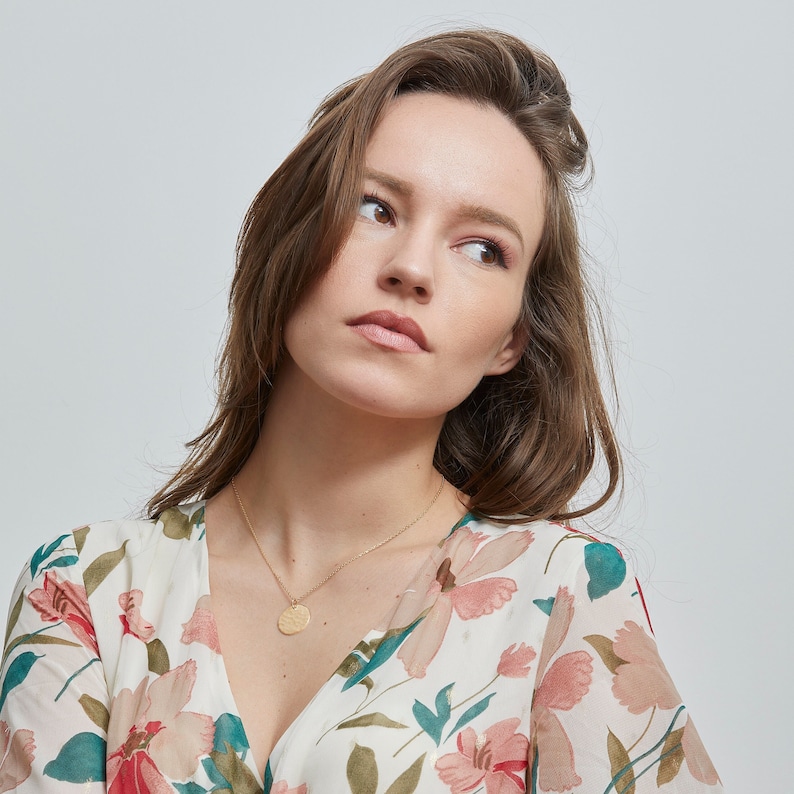 Puede incluir: Una mujer con el pelo largo y casta&ntilde;o que lleva un vestido blanco con estampado floral, un collar de oro y un colgante redondo.