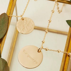 Puede incluir: Un collar de oro con dos colgantes redondos. Un colgante es texturizado y el otro tiene la inscripci&oacute;n "Te quiero" en cursiva.