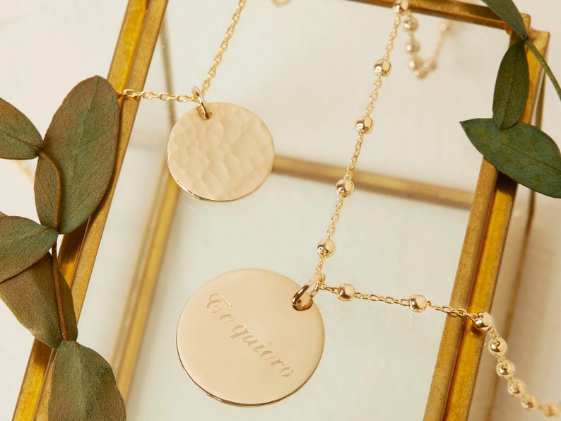 Puede incluir: Un collar de oro con dos colgantes redondos. Un colgante es texturizado y el otro tiene la inscripci&oacute;n "Te quiero" en cursiva.