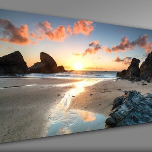 May include: A scenic sunset over a sandy beach with rocky outcrops. The sky is a vibrant mix of pink, orange, and blue with fluffy clouds. The sun is setting over the horizon, casting a golden glow on the water.
