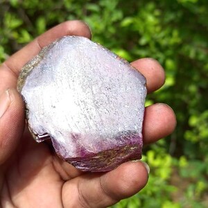 May include: A large, rough, purple and pink gemstone with a white, crystalline interior. The gemstone is being held in a hand.