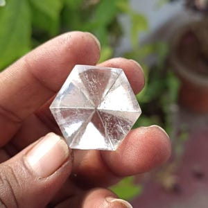 Puede incluir: Un cristal hexagonal transparente sostenido en una mano. El cristal tiene múltiples facetas que reflejan la luz, creando un efecto brillante. El fondo presenta follaje verde y una planta en maceta.