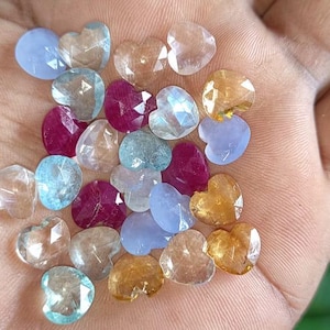 Könnte beinhalten: Eine Hand, die eine Sammlung herzförmiger Edelsteine in verschiedenen Farben hält. Die Edelsteine umfassen Farbtöne von Blau, Rosa, Klar und Gold. Die Edelsteine sind facettiert, fangen das Licht ein und erzeugen einen funkelnden Effekt. Der Hintergrund ist verschwommenes grünes Laub.