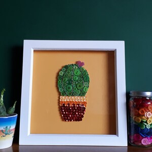 May include: A framed artwork featuring a cactus made of buttons. The cactus is green with a red and orange pot. The background is a light orange colour. The frame is white.