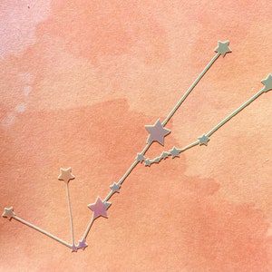 May include: A constellation of silver stars on a light orange background. The stars are connected by thin lines to form a constellation.