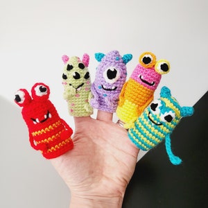 Può includere: Una mano che mostra cinque colorati burattini a dito all'uncinetto. Ogni burattino è un design di mostro unico, con colori vivaci come rosso, verde, viola, giallo e blu. Hanno varie espressioni facciali e dettagli.