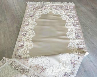 May include: Beige prayer rug with a floral design in white and purple. The rug has a decorative border and fringe on the edge. The design is symmetrical and features a central arched shape.