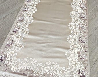 May include: A rectangular prayer rug in a soft beige color featuring a central arch design. The border is decorated with intricate white floral patterns and subtle purple accents. The rug is finished with white fringe along both short ends.