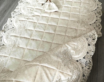 May include: A cream-colored prayer mat featuring a quilted, grid-patterned center and wide, intricate lace borders with floral embroidery. A small, matching cream-colored pouch with a drawstring and a beaded tassel rests on the mat surface.
