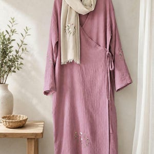 May include: A mauve wrap dress with long sleeves and a V-neckline, featuring delicate floral embroidery on the lower front and sleeves. A beige scarf with matching floral embroidery is draped over the shoulders of the dress.