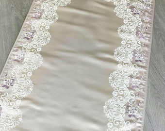 May include: A rectangular prayer rug in a soft beige satin finish, featuring an intricate white lace-like border with purple floral accents. The rug has a smooth central panel and is finished with a delicate white fringe along the top edge.