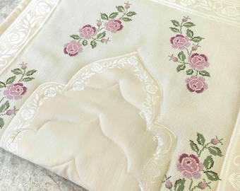 Op de afbeelding: Een crèmekleurig gebedskleed met een centraal mihrab-boogontwerp en een witte rozenrand. De stof is versierd met geborduurde bloemmotieven in roze en groene tinten, met trossen rozen en bladeren op een textuurrijk oppervlak.