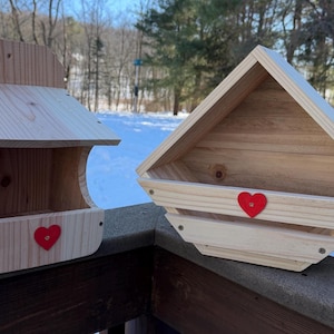 May include: Two wooden birdhouses, one rectangular and one triangular, both with natural wood tones. Each birdhouse has a red heart accent. The birdhouses are on a wooden deck with a snowy background.