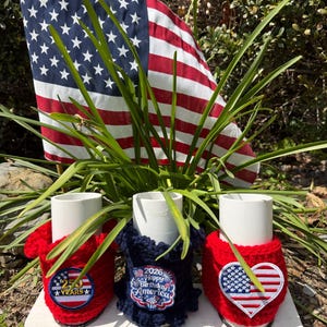 May include: Three white cylindrical tubes wrapped in textured crochet covers. The left cover is red with a circular patch reading 250 YEARS. The center is navy blue with a patch reading 2026 Happy Birthday America. The right is red with a heart-shaped flag patch.