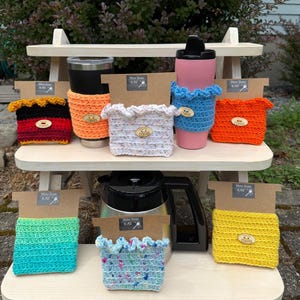 May include: A display of colorful, crocheted coffee cup sleeves and tumblers on a wooden picnic table. Sleeves are in various colors, including red, orange, blue, and yellow. Each sleeve has a wooden button that says "Coffee".