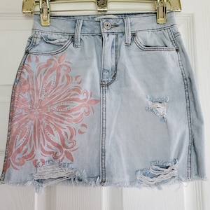 May include: Light blue denim skirt with a distressed hem and a pink floral design on the left side. The skirt has a few rips and tears.