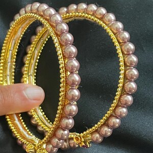 May include: Two gold bangles with a row of mauve faux pearls around each. The bangles are stacked on top of each other.