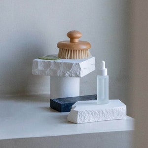 May include: White ceramic and wooden bathroom countertop decor with a brush, a bottle of serum, and a small green soap.