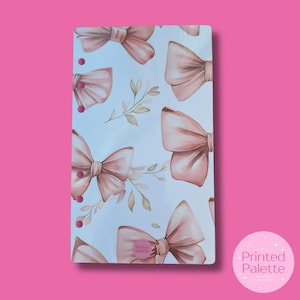 May include: A white planner cover with a repeating pattern of pink watercolor bows and leaves. The cover has six pink dots along the left side. The text "Printed Palette" is printed in a circle at the bottom of the cover.