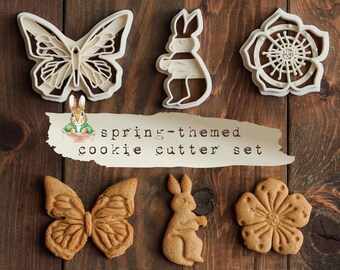 May include: A set of four spring-themed cookie cutters. The cutters are shaped like a butterfly, a rabbit, a flower, and a butterfly. The cutters are made of white plastic and are arranged on a wooden surface. The text "spring-themed cookie cutter set" is written on a piece of paper in the center of the image.